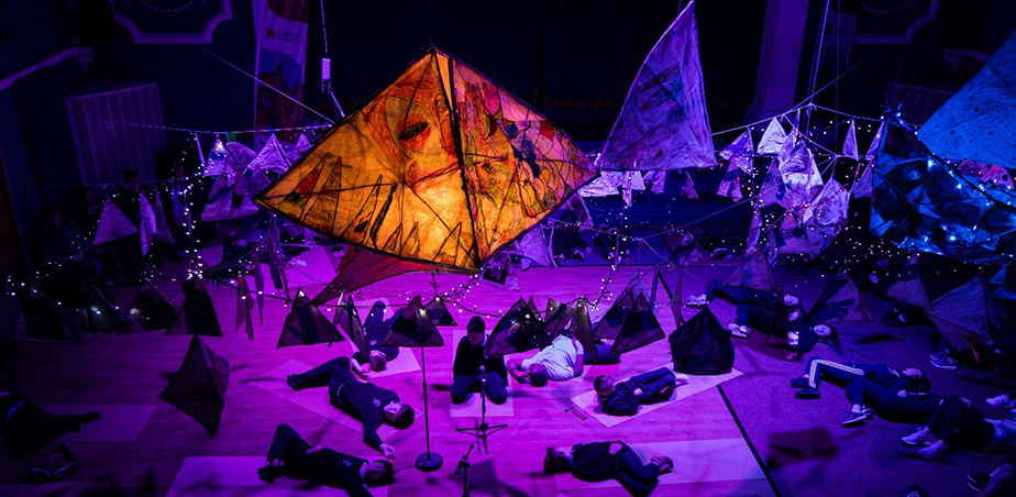 Orange paper fish sculpture hovering over purple stage with people lying down: Creative Schools Week 2019, Mullingar