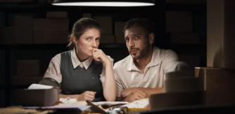 A woman and man sit at a table strewn with boxes and paraphanalia in a darkened warehouse