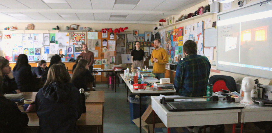 A wide shot of the interior of an art classroom with students sitting in groups at their tables listening to guest artists addressing them. 