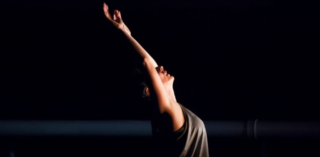 A dancer extends her arms backwards above her head. The background is dark.