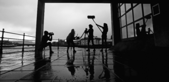 Four young people are shot in silhouette under a veranda on a pier. Each person is holding a piece of filming equipment in an active pose from left to right: camera, clapperboard, tripod, and boom microphone.