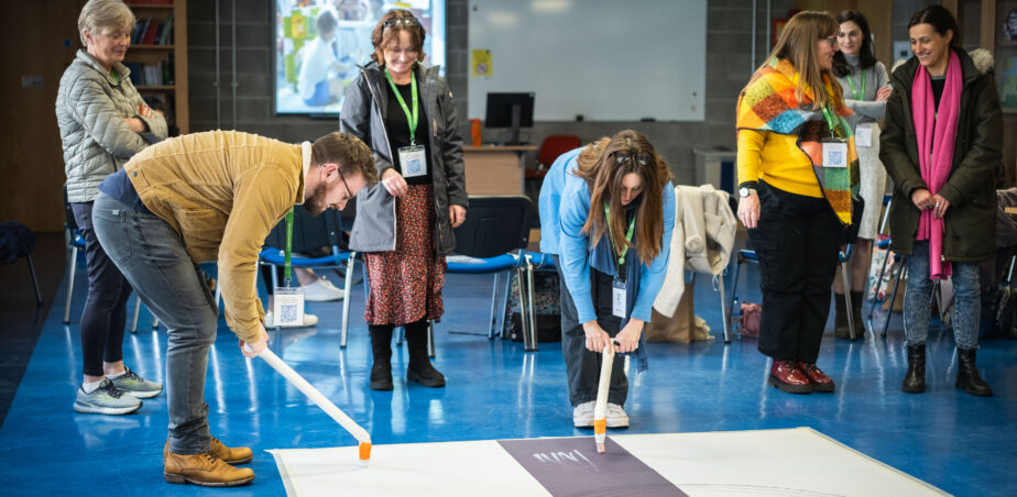 A group of adults partake in a mark making workshop drawing on a large piece of paper taped to a blue floor. Two people are bent over holding white sticks with chalk at the end drawing on the paper.