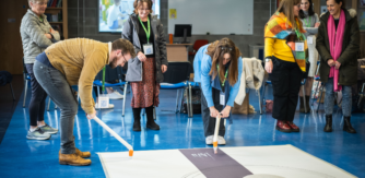 A group of adults partake in a mark making workshop drawing on a large piece of paper taped to a blue floor. Two people are bent over holding white sticks with chalk at the end drawing on the paper.