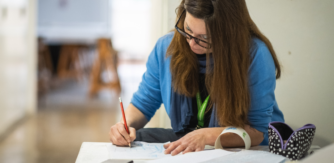 A woman in a vlue cardigan sits at a desk drawing with paper and a pencil