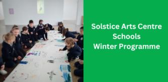 In Solstice Gallery, a large room with white walls, young children sit on the floor in two rows to the left and right of image. There is a large long sheet of white paper in-between them were they are drawing on with colourful crayons.