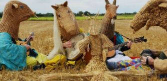 Musicians wearing straw weaved heads sit in a cut wheat field.