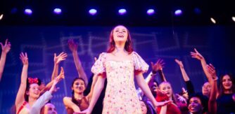 A girl performs stage front with an assemble of performers behind her