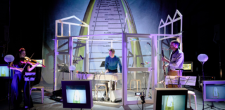 Three musician stand on a stage surrounded by screens and wires. On the left is a women playing a viola, in the middle is a main playing a keyboard and to the right another man playing a bass clarinet