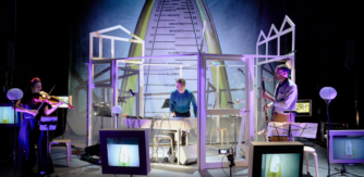 Three musician stand on a stage surrounded by screens and wires. On the left is a women playing a viola, in the middle is a main playing a keyboard and to the right another man playing a bass clarinet