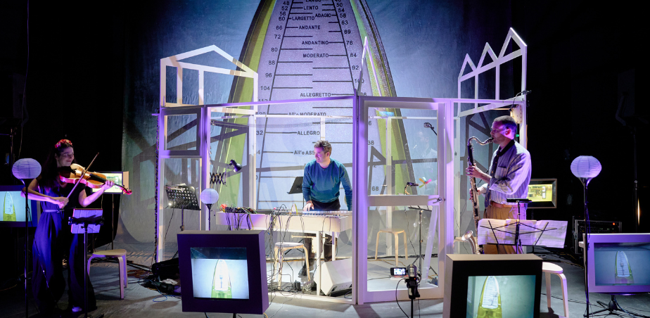 Three musician stand on a stage surrounded by screens and wires. On the left is a women playing a viola, in the middle is a main playing a keyboard and to the right another man playing a bass clarinet